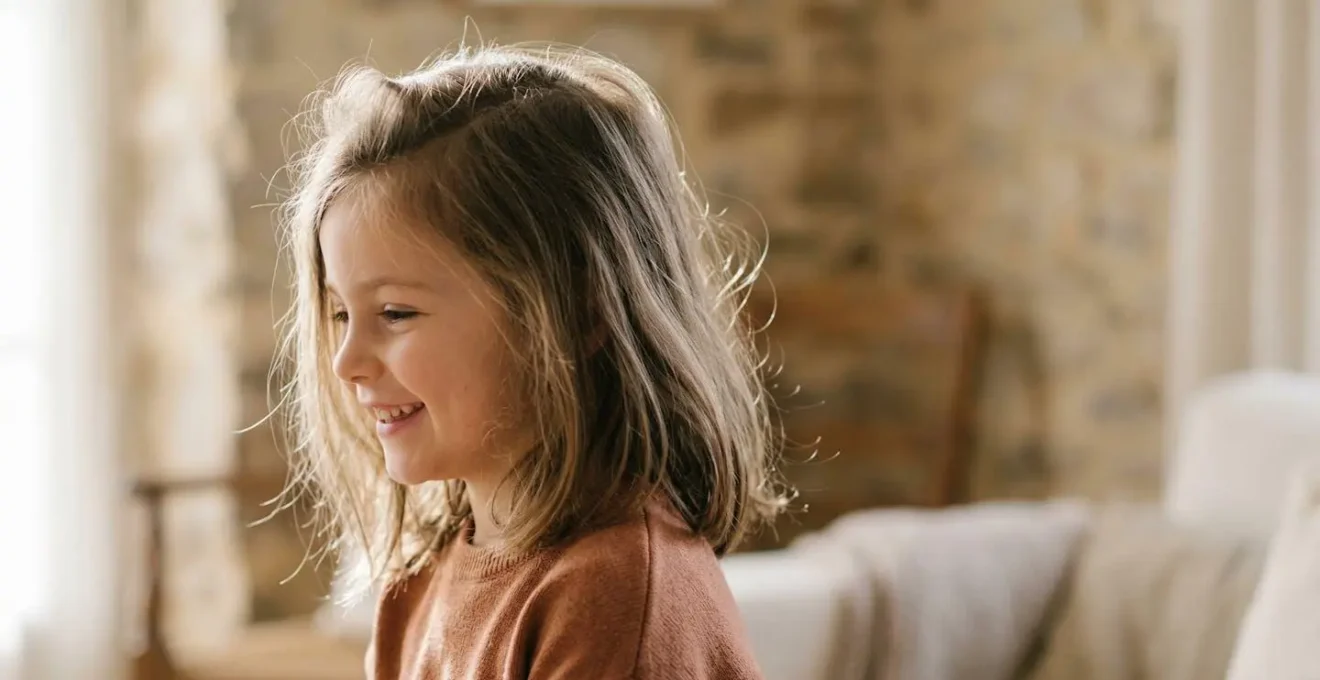 Petite fille souriante aux cheveux fins volumisés naturellement, de profil dans une lumière douce